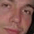 A pixelated close-up of a person's face showing eyes, nose, and mouth with a neutral expression against a light brown background.