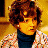 Pixelated static image of a person with curly brown hair wearing a dark turtleneck and patterned jacket against a yellow background.