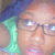 Pixelated close-up of a person with light skin wearing glasses and blue hair, holding a green object near their face.
