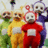Four cartoon characters in colorful full-body suits stand side-by-side against a light background with faint vertical lines.