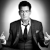 Black-and-white pixelated image of a person in a suit with a tie, seated and gesturing with both hands raised at shoulder level.