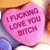 A pink heart-shaped candy with pixelated text reading "I FUCKING LOVE YOU BITCH" in dark purple letters, set against a blurred background of warm colors.