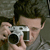 A person with short dark hair holds a silver digital camera with a black lens up to their face, positioned against a blurred background.