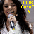 A person with long dark hair holds a microphone, wearing white clothing against a gray background with yellow text and star symbols.