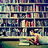 A pixelated static image shows a person sitting at a desk with multiple bookshelves filled with books in the background.