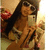 A woman with long dark hair and sunglasses takes a selfie in front of a mirror, holding a white phone, wearing a light-colored top.
