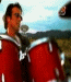 A man with dark hair and sunglasses plays red drums in a field under a cloudy sky, captured across showing head movement