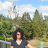 Pixelated outdoor scene with a person in dark clothing standing in front of green trees and a tall white structure under a blue sky with clouds.