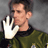 Pixelated static image of a person in profile wearing a green jacket with black collar, holding up a white hand gesture against gray background.