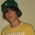 A person wearing a dark green cap and yellow shirt with partially visible text on the chest, set against a light brown background.