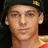Pixelated image of a person wearing a dark cap with yellow emblem, red shirt, and earrings against blue background with pink text.
