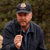 Pixelated image of a person wearing a dark cap with a light circular patch, dark jacket, and light-colored shirt, holding a small object near their chest.
