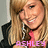 A pixelated image of a smiling person with long blonde hair wearing a black jacket over a pink top, with the name 'ASHLEY' displayed in stylized text below.