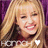 Pixelated image of a smiling person with long blonde hair against a purple background, wearing a dark top with a red accent and a white heart-shaped symbol.