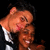Two figures in a close embrace, one with light brown hair and the other with dark hair, against a solid black background.