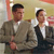 Two men in formal attire stand side-by-side against a blurred background with warm tones.
