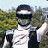 Pixelated image of a person wearing a dark cap with a white stripe, white shirt, and dark vest, standing outdoors with blurred greenery in the background.