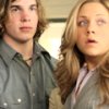 Two people with long light brown hair sit side-by-side in a vehicle interior, wearing gray button-up shirts with visible buttons.