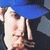 A static pixelated image shows a person with light skin wearing a blue cap and white shirt, holding an object near their face against a dark background.