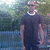Pixelated static image of a person in dark clothing standing outdoors with blurred greenery and water background.