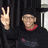 Pixelated image of a person making a peace sign with their right hand, wearing a dark top and red collar against a black background.