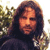 A pixelated portrait of a person with long dark hair wearing a dark top, set against a blurred background with green and yellow tones.