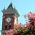 Pixelated image of a red brick clock tower with a gray roof and clock face, surrounded by pink flowering trees under a light blue sky.