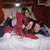 A group of people in a bed with white sheets, one person in red clothing leaning over another person, and a third person lying down.
