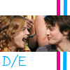 Two people with curly hair face each other closely, one with mouth open in laughter, against a background with vertical pink and blue stripes and blue text 'D/E' at bottom left.
