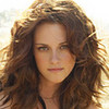 A static image of a person with long wavy brown hair, green eyes, and a neutral expression, wearing a dark strap top against a bright background.