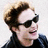 A person with light brown curly hair and dark sunglasses wears a black jacket, smiling while turning head left to right across four