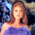 Static 50x50 pixel buddy icon showing a woman with light brown hair in a purple top against a blurred background with blue and beige tones.