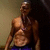 A shirtless man with a muscular build stands against a dark background, wearing blue shorts and a silver chain necklace, with his head turning slightly left to right across frames.