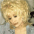 Pixelated close-up of a person with light brown hair styled in a voluminous updo, wearing a light-colored top against a muted background.