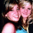 Two women in close pose, one with brown hair and blue top, the other with light brown hair; both smiling against dark background.