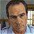 A pixelated close-up of a person with short dark hair wearing a light blue shirt, set against a blurred background with a window and furniture.