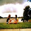 Two people sit on grass near a body of water, holding hands under a cloudy sky with trees in the background.