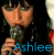 A 50x50 pixel animated buddy icon showing a person holding a microphone with the name "Ashlee" displayed in blue text on the left frame and white text on the right frame, arranged as a contact sheet of two frames.