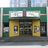 Static image of a building facade with green and yellow sections, two white rectangular signs above the entrance, and a dark doorway flanked by yellow pillars.