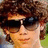 Pixelated close-up of a person with curly brown hair wearing dark sunglasses and a light-colored shirt against a blurred background.