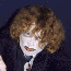 A person with curly brown hair and pale makeup wears a dark robe over a white shirt and dark tie, performing sequential hand and head movements against a dark blue background.