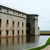 Pixelated static image of a light-colored building with dark roof and columns, partially reflected in water, with green grass and a distant horizon visible.