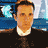 Pixelated static image of a man with light brown hair and orange skin tone wearing a white collared shirt under a dark jacket, set against a blue background with abstract shapes.