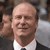 A pixelated static image of a bald man in a dark suit and white shirt, smiling with closed eyes and a slight head tilt, against a blurred background.