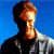 Pixelated profile of a person with short dark hair against a solid blue background, wearing a dark top with light-colored shoulder accents.