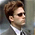 Pixelated close-up of a person with brown hair wearing dark sunglasses, a white collared shirt, and a dark suit jacket against a blurred background.