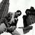 Pixelated grayscale image of a figure holding a microphone with another figure partially visible behind, set against a plain background.