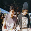 Two people stand side-by-side under a dark canopy; one wears a gray beanie and a dark shirt with a light graphic, the other a light gray shirt with a faint design.