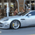 A silver sedan with a visible wheel and yellow hubcap is positioned on a street, with blurred figures and buildings in the background.