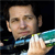 A pixelated close-up of a person wearing a dark jacket with blue and green accents, holding a light-colored object against a dark background.